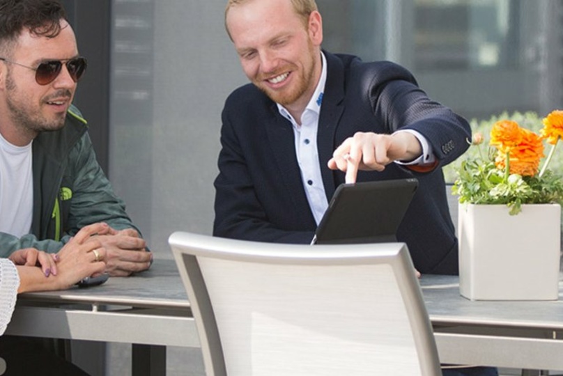 Drei Personen sitzen an einem Tisch im Freien, ein Mann zeigt auf ein Tablet.